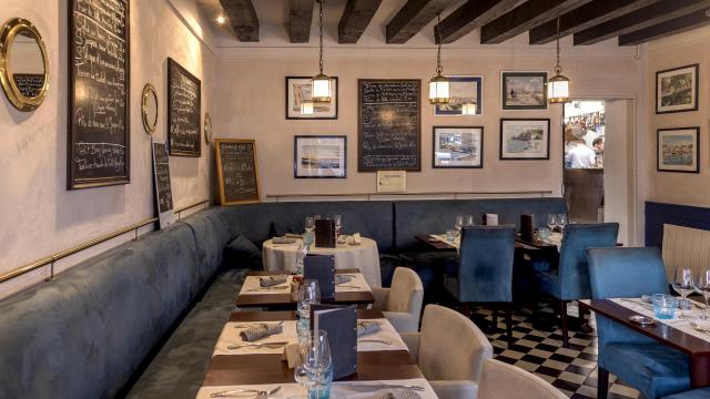 Salle restaurant d'un bistrot dieppois, avec de nombreux cadres et ardoises au mur, tables dressées autour de banquettes et chaises en velours bleus, sol en damier noir et blanc. On aperçoit l'effervescence dans la cuisine à travers la porte ouverte.