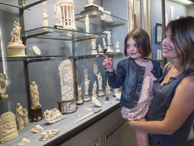 Une maman porte sa fille dans les bras pour qu'elle puisse admirer une vitrine présentant des sculptures en ivoire