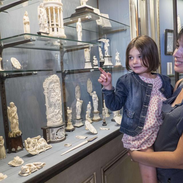 Une maman porte sa fille dans les bras pour qu'elle puisse admirer une vitrine présentant des sculptures en ivoire