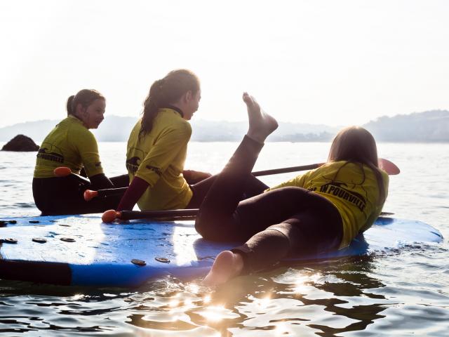 3 jeunes filles assises ou allongées sur des paddles devant un coucher de soleil