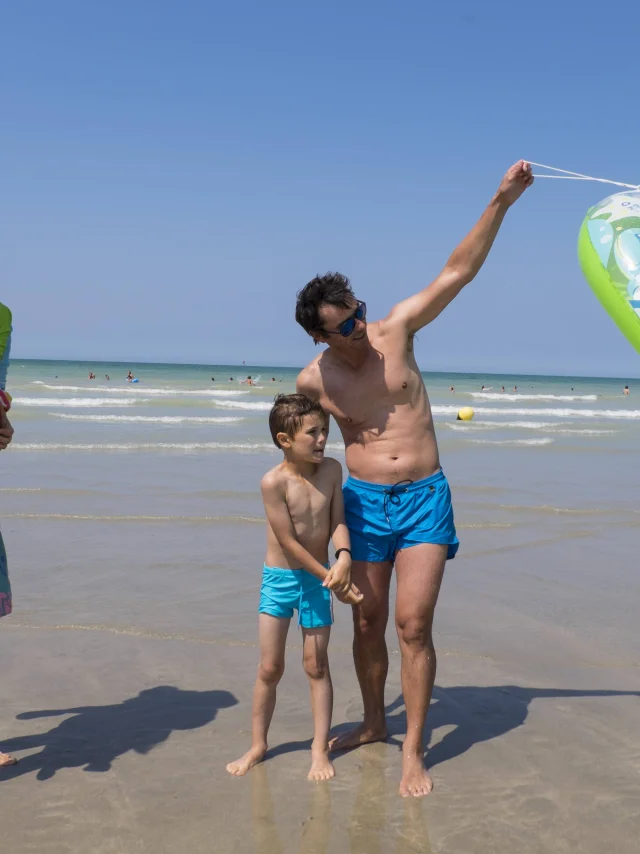 Famille de 4 personnes en maillot de bain sur la plage de Pourville à marée basse. Le papa soulève une bouée.