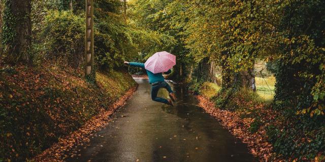 Homme de dos sautant avec un parapluie rose dans la rue de campagne bordée d'arbres aux couleurs automnales