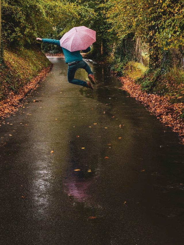 Homme de dos sautant avec un parapluie rose dans la rue de campagne bordée d'arbres aux couleurs automnales