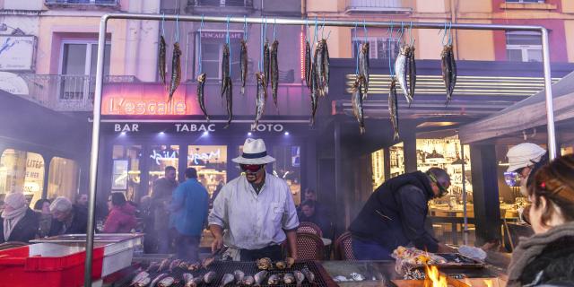 Homme faisant griller des harengs devant le bar l'Escale sur le quai Henri 4. Des harengs crus sont accrochés à une barre au-dessus du grill. A l'arrière-plan des personnes sont attablées en terrasse