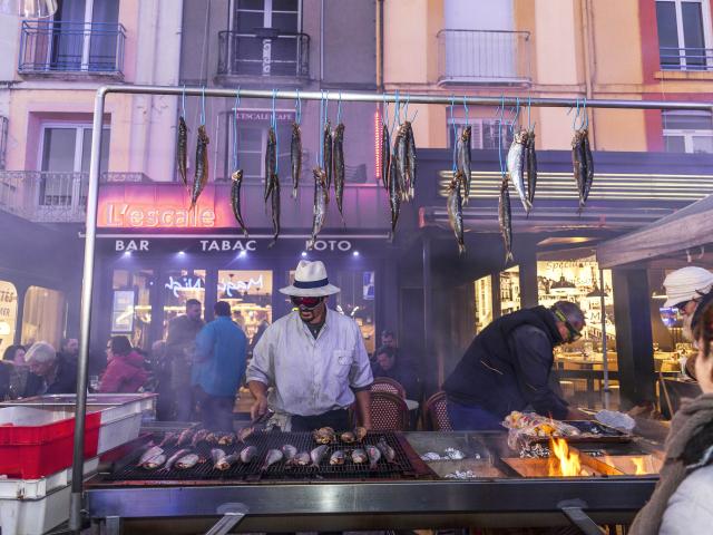 Homme faisant griller des harengs devant le bar l'Escale sur le quai Henri 4. Des harengs crus sont accrochés à une barre au-dessus du grill. A l'arrière-plan des personnes sont attablées en terrasse