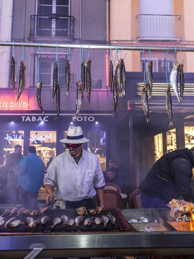 Homme faisant griller des harengs devant le bar l'Escale sur le quai Henri 4. Des harengs crus sont accrochés à une barre au-dessus du grill. A l'arrière-plan des personnes sont attablées en terrasse