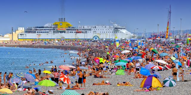 Plage de Dieppe en plein été. Nombreuses personnes installées sur les galets avec des parasols de toutes les couleurs. Le ferry entre dans le port en arrière-plan.