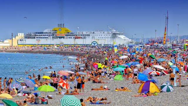 Plage de Dieppe en plein été. Nombreuses personnes installées sur les galets avec des parasols de toutes les couleurs. Le ferry entre dans le port en arrière-plan.