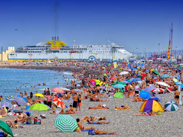 Plage de Dieppe en plein été. Nombreuses personnes installées sur les galets avec des parasols de toutes les couleurs. Le ferry entre dans le port en arrière-plan.