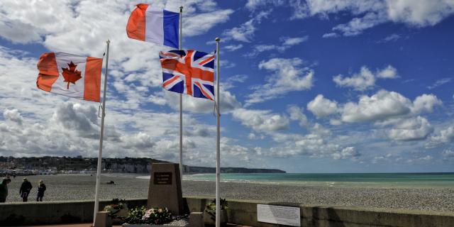 Drapeaux canadien, français et anglais flottant autour d'une stèle commémorative, plage et falaises en arrière-plan