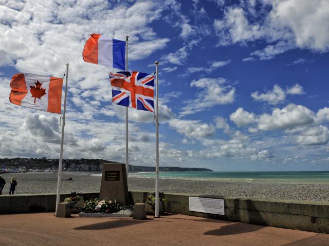 Drapeaux canadien, français et anglais flottant autour d'une stèle commémorative, plage et falaises en arrière-plan