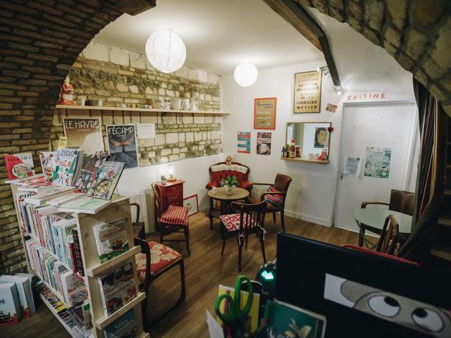 Coin goûter dans une librairie. Pièce voûtée, fauteuils dépareillés, affiches aux murs, étagère remplie de livres.