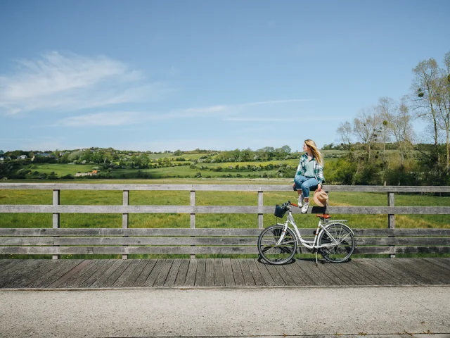 Une jeune fille est assise sur une rembarde en bois, son vélo devant elle. A l'arrière-plan, le paysage verdoyant de la Vallée de la Scie.