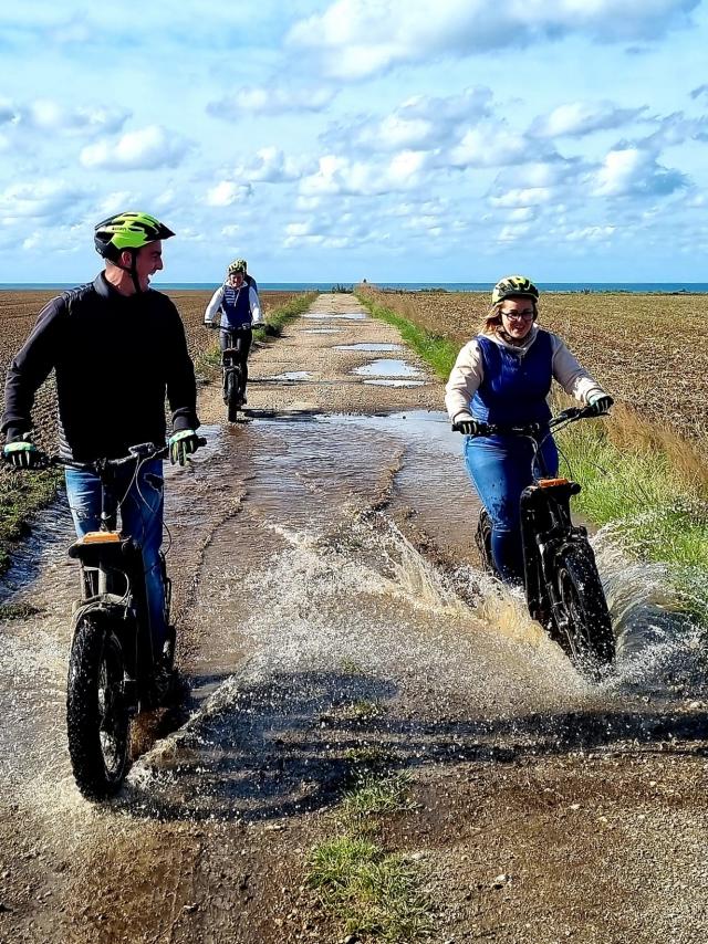 3 personnes font de la trottinette électrique tout terrain sur un sentier. On aperçoit la mer au loin et un participant roule dans une flaque d'eau qui éclabousse la jeune femme à côté.