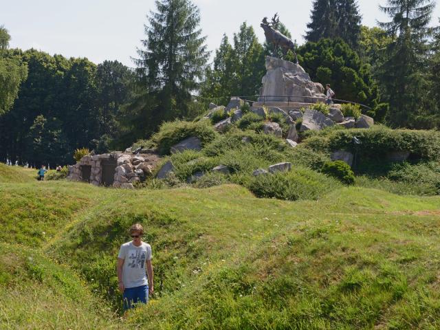 Un caribou en bronze au sommet d'un promontoire rocheux au milieu d'un espace forestier et d'anciennes tranchées aujourd'hui recouverte d'herbe.