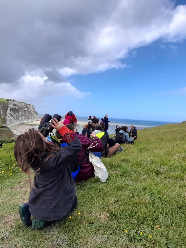 Un groupe d'enfants et d'adultes est assis sur la falaise au-dessus de la plage de Petit-Caux. Une petite fille regarde le ciel avec des jumelles.