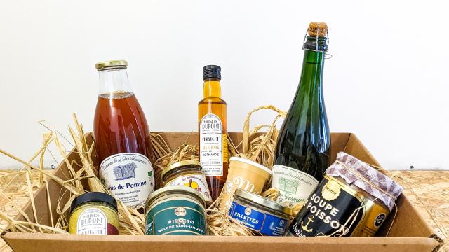 Une boîte en carton avec de la paille à l'intérieur de laquelle sont disposées des produits locaux de la région dieppoise : jus de pomme, vinaigre de cidre, cidre fermier, caramel de pommes, miel, moutarde, risotto de Saint-Jacques, rillettes de maquereau, soupe de poisson