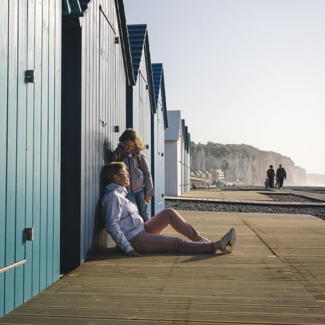 Une maman et sa fille sont assises devant une cabine de plage bleue à Dieppe et rient ensemble, avec les falaises et des promeneurs en arrière-plan