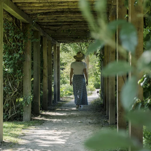 Une femme de dos traverse un couloir en bois dans un jardin verdoyant