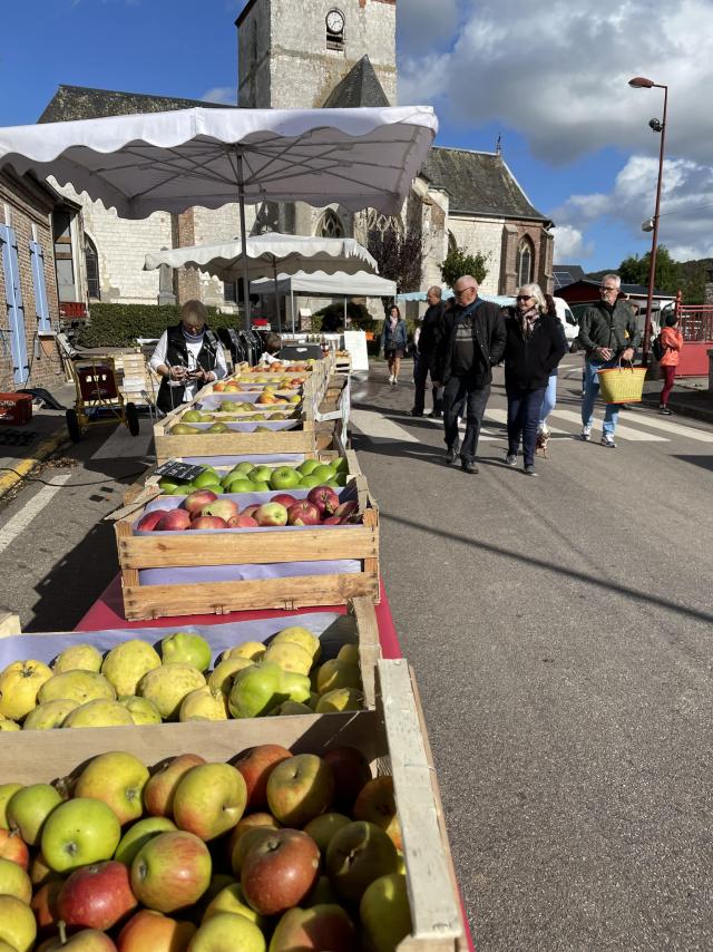 Une exposante de pommes dans des cagettes et des passants devant l'église d'un village normand