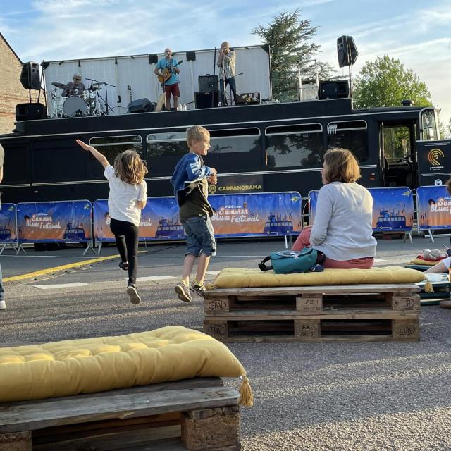 Des enfants en train de danser et un public assis sur des banquettes faites en palette devant un groupe de musique qui joue sur le toit d'un bus aménagé