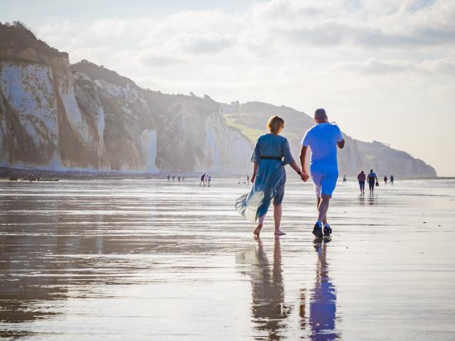 Un couple homme et femme se balade main dans la main sur la plage de Pourville à marée basse, falaises en arrière plan