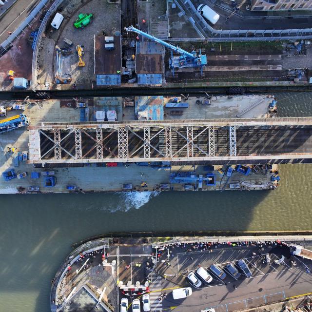 Un grand pont en fer de style Eiffel en travaux sur la barge qui le transporte dans le port.