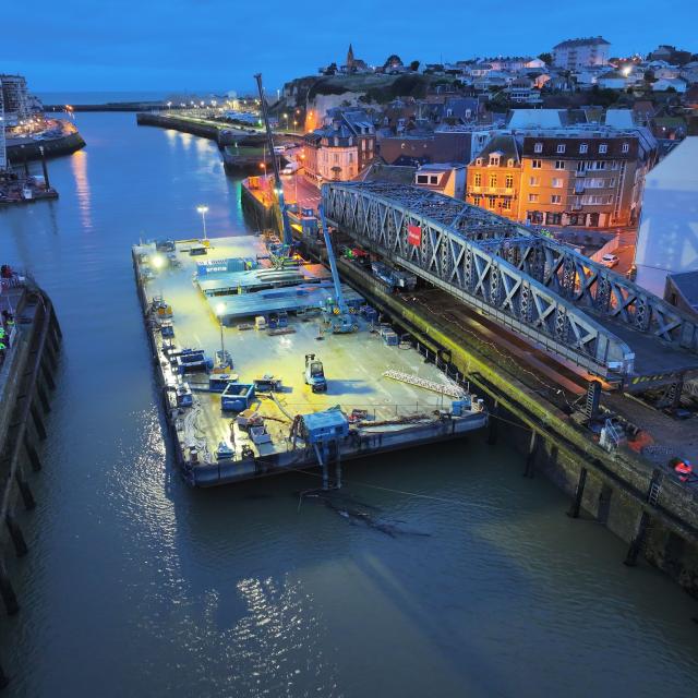 A la tombée de la nuit, un grand pont en fer de style Eiffel en travaux, à côté de la barge qui doit le transporter dans le port