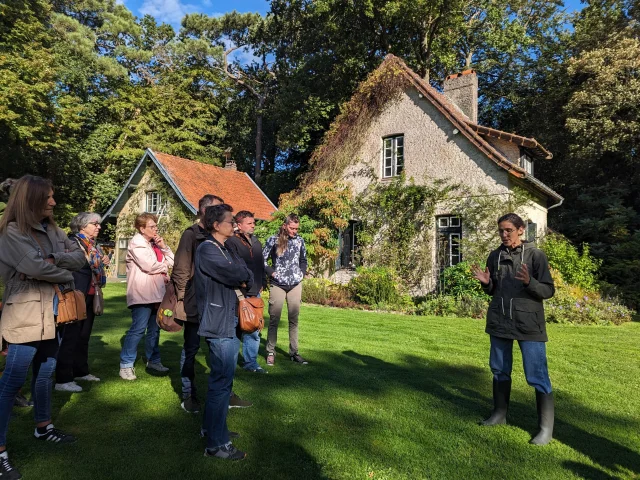 Groupe des personnes écoutant une guide conférencière dans un jardin fleuri avec une maison en arrière plan